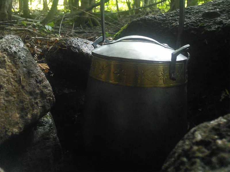 A kettle with a bright engraved band hanging beneath a rock overhang, woods behind